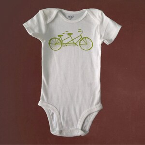 May include: White baby onesie with a green tandem bicycle graphic. The onesie has a snap closure at the bottom.