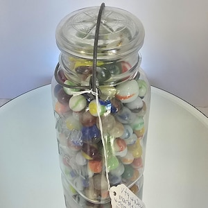 May include: A clear glass jar filled with a variety of colorful marbles. The jar has a metal handle and a glass lid. A small tag is attached to the jar. The marbles are in shades of red, blue, green, yellow, and white.