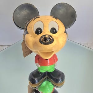 May include: A vintage Mickey Mouse figurine featuring a large, tan-colored head with black ears, a red shirt, bright green pants, and black shoes. The character has a wide, open-mouthed expression and a worn black nose, with a small brown tag attached.