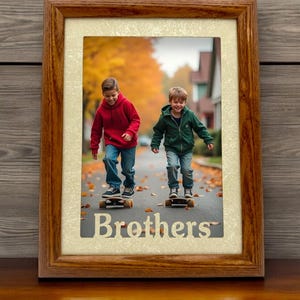 May include: A framed photo of two brothers skateboarding down a street lined with autumn leaves. The photo is in a brown wooden frame with a cream-colored mat. The word "Brothers" is printed in gold lettering below the photo.