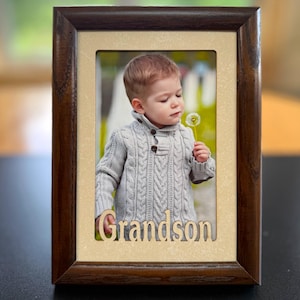 May include: A wooden picture frame with a photo of a young child holding a dandelion. The frame has a dark brown finish with a light beige mat. The word "Grandson" is cut out at the bottom of the frame.