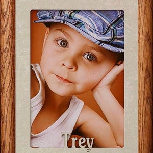 May include: A framed photo of a young boy wearing a blue and white striped hat. The boy is looking at the camera with a thoughtful expression. The frame is made of wood and has a light brown finish. The boy's name, "Trey", is written in silver letters below the photo.