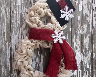 May include: A snowman-shaped wreath made of beige burlap, with a burgundy scarf and black top hat. White snowflake accents are attached to the hat and scarf. A black ribbon is used for hanging. The wreath is displayed against a weathered, white-painted wooden background.