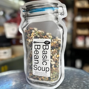 May include: A clear, jar-shaped bag filled with a mix of dried beans. The bag has a blue and silver closure and a label that reads "Basic Bean Soup" with "GF" for gluten-free. The label also includes the text "Made in the USA with Pride".