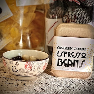 May include: A small ceramic bowl with a red and black floral design holds dark chocolate-covered espresso beans and light-colored candies. A package of chocolate-covered espresso beans is in the background, with the text "CHOCOLATE COVERED ESPRESSO BEANS".