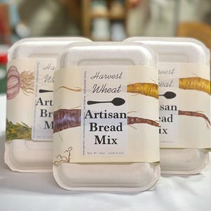 May include: Three rectangular containers of Artisan Bread Mix, each wrapped with a label featuring the text "Harvest Wheat" and "Artisan Bread Mix." The labels display illustrations of garlic, herbs, and root vegetables.
