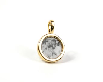 Può includere: Un ciondolo rotondo color oro con una piccola fotografia in bianco e nero all'interno. Il ciondolo ha un anello nella parte superiore per una catena. La fotografia mostra due persone sorridenti. Il ciondolo è su sfondo bianco.