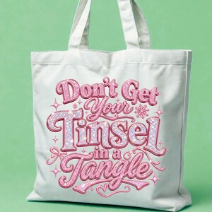 May include: A white canvas tote bag featuring a pink glittery graphic print with the text "Don't Get Your Tinsel in a Tangle" surrounded by small star and snowflake accents. The bag has two top handles and a rectangular shape.