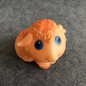May include: A small, orange rubber toy sheep with blue eyes. The sheep has a fluffy texture and a smiling expression.