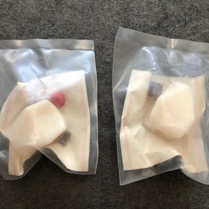 Op de afbeelding: Twee doorzichtige plastic zakken met witte, ronde, gladde voorwerpen. Eén zak bevat ook een klein rood voorwerp en een klein zwart voorwerp. De andere zak bevat een klein zwart voorwerp.