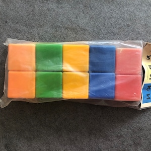 Può includere: Un set di 12 blocchi di plastica colorati in una busta di plastica trasparente. I blocchi sono arancioni, verdi, gialli, blu e rossi. La busta ha un'etichetta che dice "KUM"