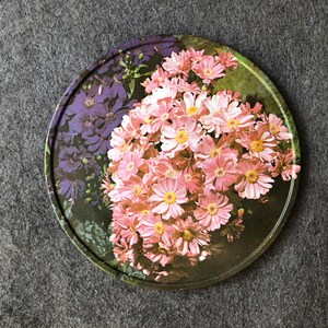 May include: A round, decorative plate with a floral design. The plate features a cluster of pink flowers with yellow centers, surrounded by green leaves. There are also some purple flowers in the background.