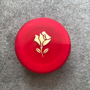 Puede incluir: Un recipiente redondo rojo con un diseño de rosa dorada en la tapa.