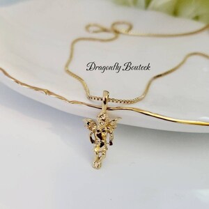 Puede incluir: Un collar de cadena de oro con un peque&ntilde;o colgante de querub&iacute;n de oro. El colgante est&aacute; detallado con alas y una pose de pie. El collar est&aacute; sobre una superficie blanca.