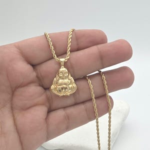 May include: A gold-toned pendant shaped like a seated figure with a rounded belly and smooth features, suspended from a matching gold-toned rope chain. The pendant has a polished, reflective surface and is attached to the chain by a small bail.