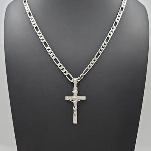 May include: A silver-colored necklace featuring a crucifix pendant. The pendant depicts a figure on a cross. The chain is a curb link style. The necklace is displayed against a black background.