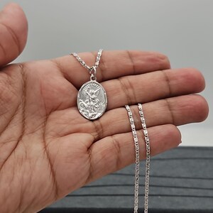 Puede incluir: Collar con colgante ovalado plateado que representa un &aacute;ngel. El colgante cuelga de una fina cadena. El collar se muestra sobre un fondo neutro.