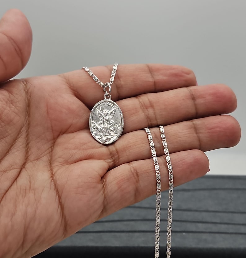 Puede incluir: Collar con colgante ovalado plateado que representa un &aacute;ngel. El colgante cuelga de una fina cadena. El collar se muestra sobre un fondo neutro.