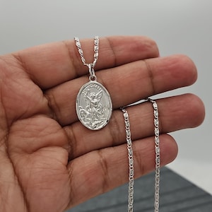 Puede incluir: Collar con colgante ovalado plateado con un &aacute;ngel grabado. El colgante cuelga de una cadena delicada. El collar se muestra sobre un fondo neutro.