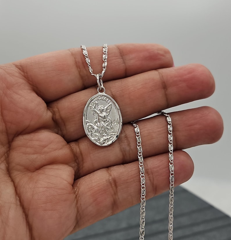 Puede incluir: Collar con colgante ovalado plateado con un &aacute;ngel grabado. El colgante cuelga de una cadena delicada. El collar se muestra sobre un fondo neutro.