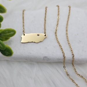 May include: Gold necklace featuring a pendant shaped like the outline of Puerto Rico with a small heart cutout. The chain is a delicate, linked style.
