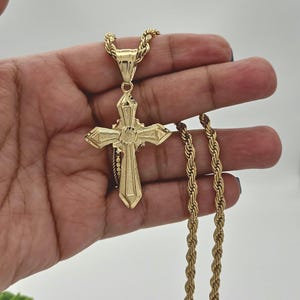 May include: A gold-tone rope chain necklace with a large, ornate gold cross pendant. The cross has a detailed design and a raised center.