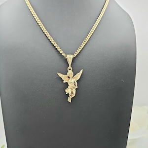 K&ouml;nnte beinhalten: Eine goldfarbene Halskette mit einem detaillierten Engel-Anh&auml;nger. Der Engel hat ausgebreitete Fl&uuml;gel und h&auml;ngt an einer Panzerkette. Die Halskette wird auf einer schwarzen Schaufensterpuppe pr&auml;sentiert, die das Design und die Farbe des Schmucks hervorhebt.