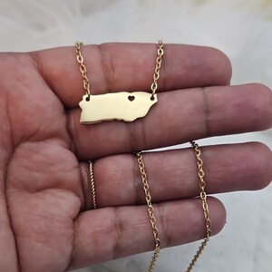 May include: Gold necklace featuring a pendant shaped like the outline of Puerto Rico with two small heart cutouts. The chain is a delicate, thin, gold chain.