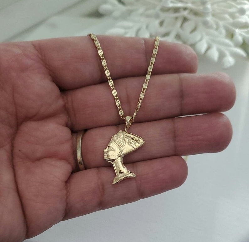 May include: A gold chain necklace with a pendant featuring a profile of an Egyptian queen wearing a headdress.