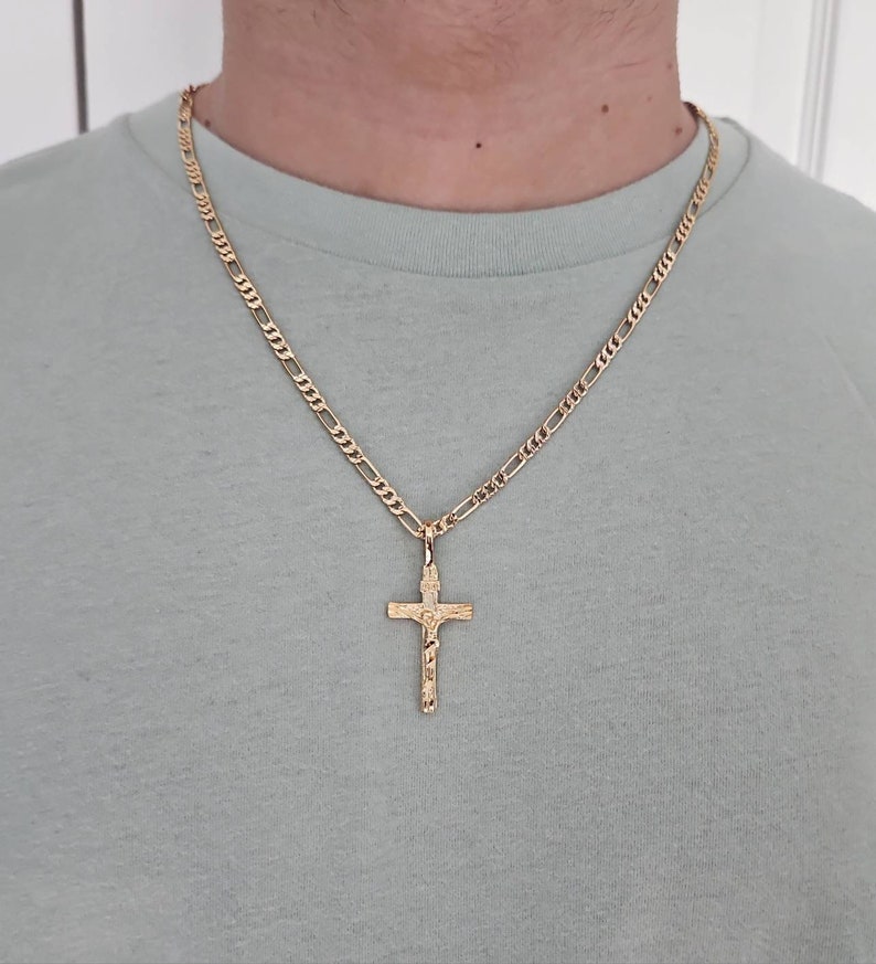 Puede incluir: Un collar de cadena de oro con un colgante de cruz de oro. La cadena es de estilo figaro.