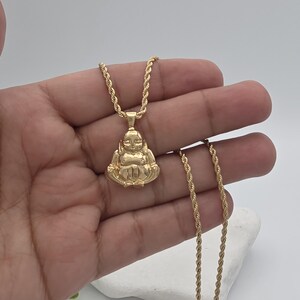 May include: A gold-toned pendant shaped like a seated figure with a smooth, polished finish, suspended from a matching gold-toned rope chain. The pendant features a stylized, rounded form with defined facial features and crossed legs in a seated position.