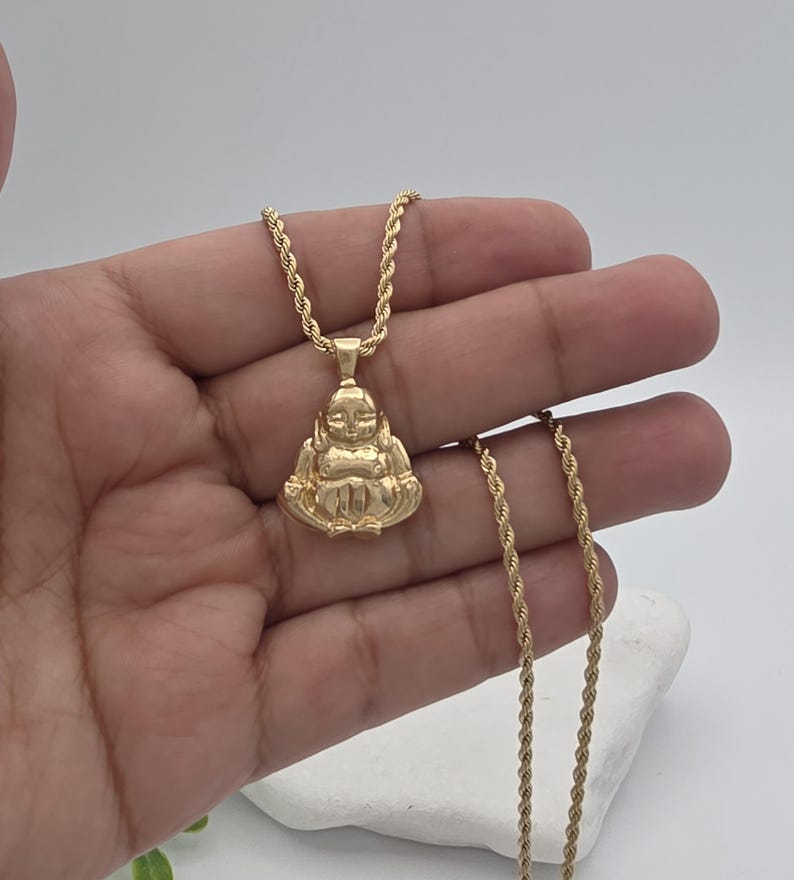 May include: A gold-toned pendant shaped like a seated figure with a smooth, polished finish, suspended from a matching gold-toned rope chain. The pendant features a stylized, rounded form with defined facial features and crossed legs in a seated position.