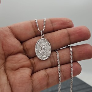 Puede incluir: Un collar con colgante plateado con un colgante ovalado grabado con la imagen de un &aacute;ngel. El colgante cuelga de una delicada cadena. El collar se muestra sobre un fondo neutro.
