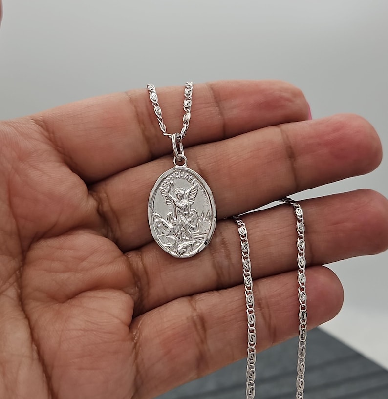 Puede incluir: Un collar con colgante plateado con un colgante ovalado grabado con la imagen de un &aacute;ngel. El colgante cuelga de una delicada cadena. El collar se muestra sobre un fondo neutro.