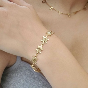 May include: A gold-colored bracelet featuring a series of turtle charms. The bracelet is worn on a wrist, and a gold necklace with the name "JAYLN" is also visible. The jewelry is set against a neutral background.