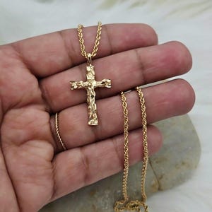 May include: A gold-colored cross pendant necklace. The cross features a textured design with a figure of Jesus Christ. The necklace chain is a rope style. The jewelry is displayed on a light-colored surface.