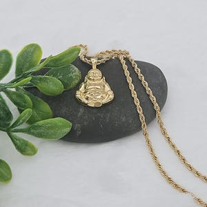 May include: A gold-toned pendant shaped like a seated Buddha figure, suspended from a matching gold-toned rope chain. The pendant features a polished, reflective surface with detailed facial features and crossed legs, resting against a dark gray stone.