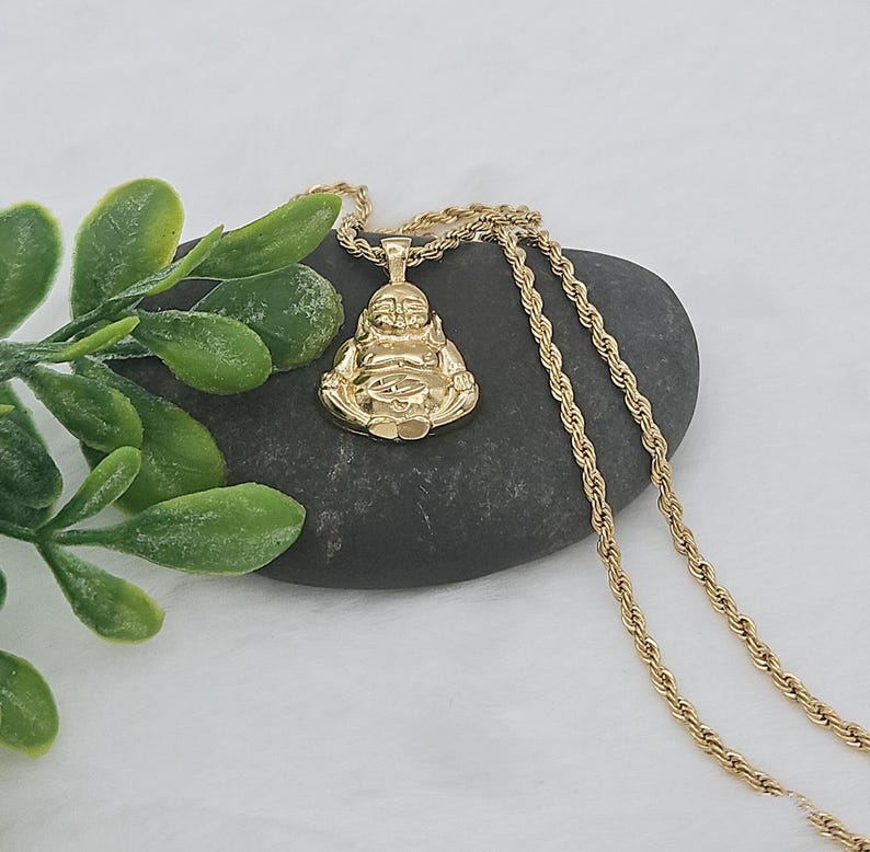May include: A gold-toned pendant shaped like a seated Buddha figure, suspended from a matching gold-toned rope chain. The pendant features a polished, reflective surface with detailed facial features and crossed legs, resting against a dark gray stone.