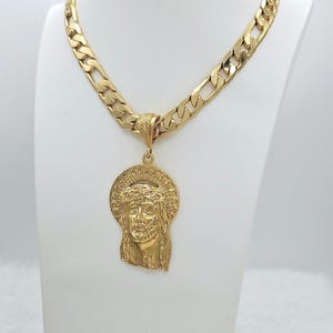 Puede incluir: Un collar dorado con una cadena gruesa y un colgante que representa a Jesús con un halo. El colgante tiene un diseño intrincado. El collar se exhibe en un busto blanco.