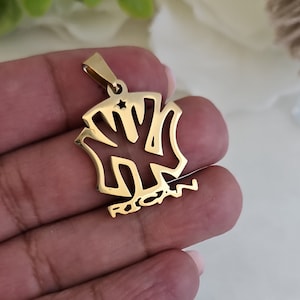 Op de afbeelding: Gouden ketting met hanger met een gestileerd 'NY'-ontwerp met een kleine ster in het midden en het woord 'RICAN' eronder.