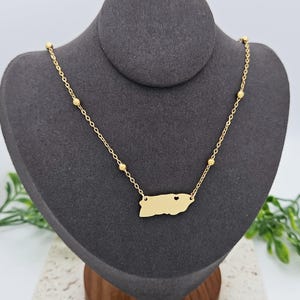 May include: Gold necklace with a small, detailed outline of Puerto Rico with a heart cutout in the center. The necklace has small gold beads along the chain.