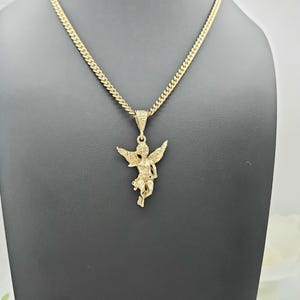 K&ouml;nnte beinhalten: Eine goldfarbene Anh&auml;ngerkette mit einem detaillierten Cherub-Design. Der Cherub hat ausgebreitete Fl&uuml;gel und h&auml;ngt an einer Panzerkette. Die Halskette wird auf einer schwarzen Schaufensterpuppe pr&auml;sentiert, die das Design und die Farbe des Schmucks hervorhebt.