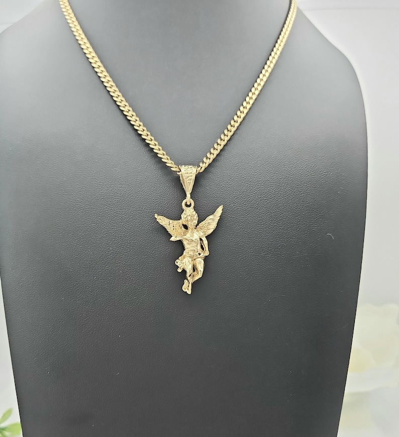 K&ouml;nnte beinhalten: Eine goldfarbene Anh&auml;ngerkette mit einem detaillierten Cherub-Design. Der Cherub hat ausgebreitete Fl&uuml;gel und h&auml;ngt an einer Panzerkette. Die Halskette wird auf einer schwarzen Schaufensterpuppe pr&auml;sentiert, die das Design und die Farbe des Schmucks hervorhebt.