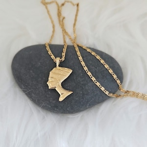 May include: A gold chain necklace with a pendant featuring a profile of an Egyptian queen. The pendant is detailed with intricate designs and is set against a gray stone background.