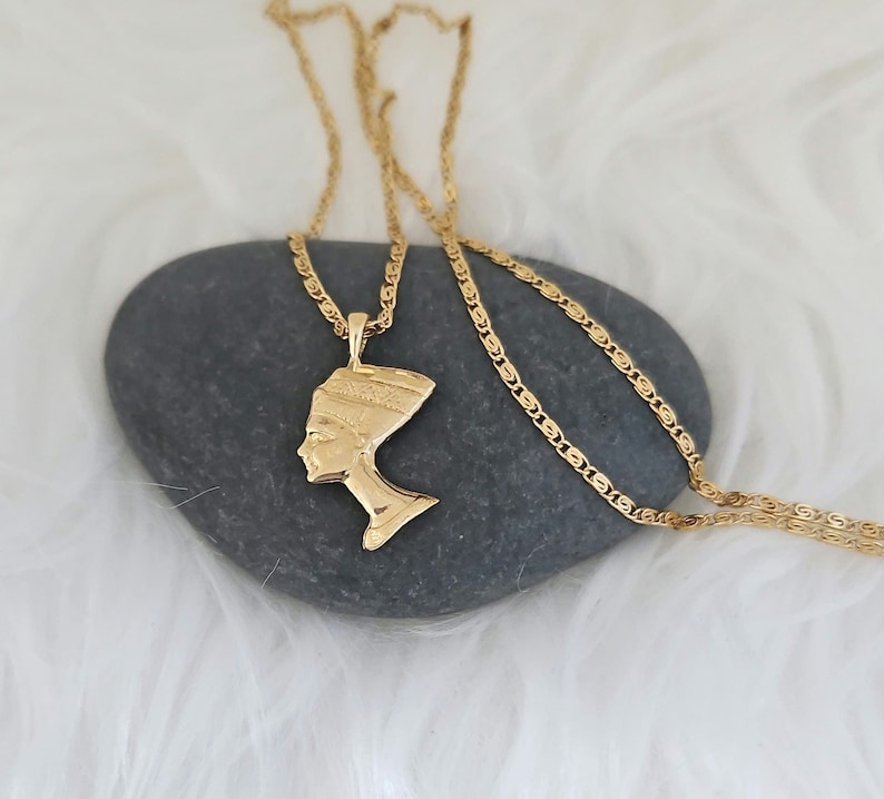 May include: A gold chain necklace with a pendant featuring a profile of an Egyptian queen. The pendant is detailed with intricate designs and is set against a gray stone background.