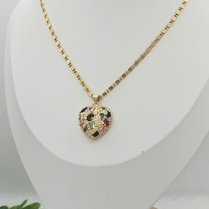 May include: A gold-colored necklace with a heart-shaped pendant. The pendant is encrusted with a variety of colorful gemstones. The necklace is displayed on a white mannequin bust.