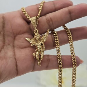 K&ouml;nnte beinhalten: Goldfarbene Halskette mit einem detaillierten Engel-Anh&auml;nger mit ausgebreiteten Fl&uuml;geln. Der Anh&auml;nger h&auml;ngt an einer dicken, strukturierten Kette. Kette und Anh&auml;nger haben eine helle, polierte Oberfl&auml;che. Die Halskette ist ein klassisches Schmuckst&uuml;ck.