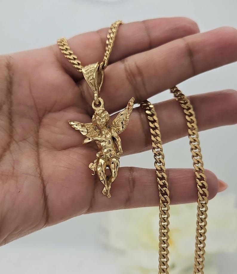K&ouml;nnte beinhalten: Goldfarbene Halskette mit einem detaillierten Engel-Anh&auml;nger mit ausgebreiteten Fl&uuml;geln. Der Anh&auml;nger h&auml;ngt an einer dicken, strukturierten Kette. Kette und Anh&auml;nger haben eine helle, polierte Oberfl&auml;che. Die Halskette ist ein klassisches Schmuckst&uuml;ck.