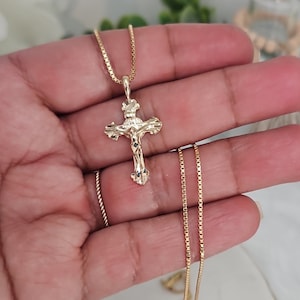 May include: A gold-colored crucifix pendant necklace. The pendant features a detailed depiction of Jesus on the cross. The necklace has a delicate chain. The jewelry is held in a hand.