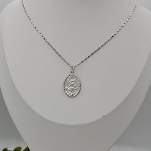 Puede incluir: Un collar plateado con un colgante ovalado grabado. El colgante cuelga de una cadena fina. El collar se exhibe en un maniqu&iacute; blanco, mostrando sus detalles.